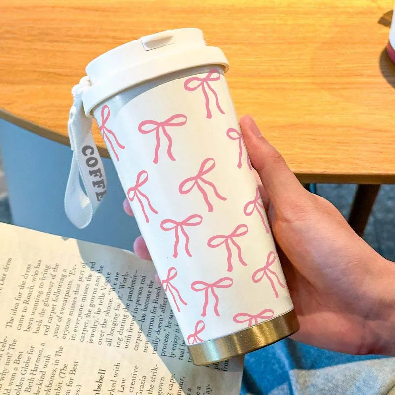 A hand holds a white travel mug with pink ribbon pattern on a wooden table.