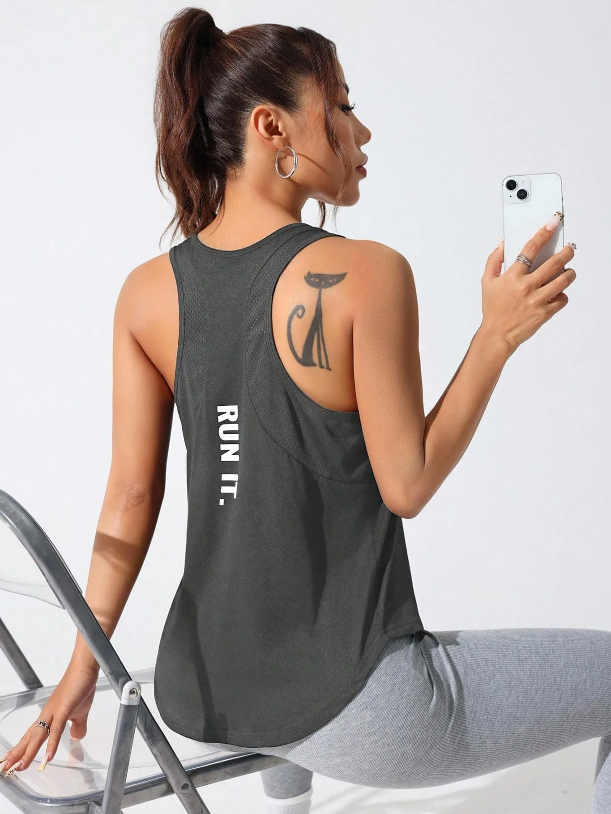 A woman wearing a dark gray running and yoga vest with a stylized letter design, sitting on a chair.