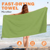 A woman holds up a large olive green microfiber quick-dry beach towel near the ocean water.