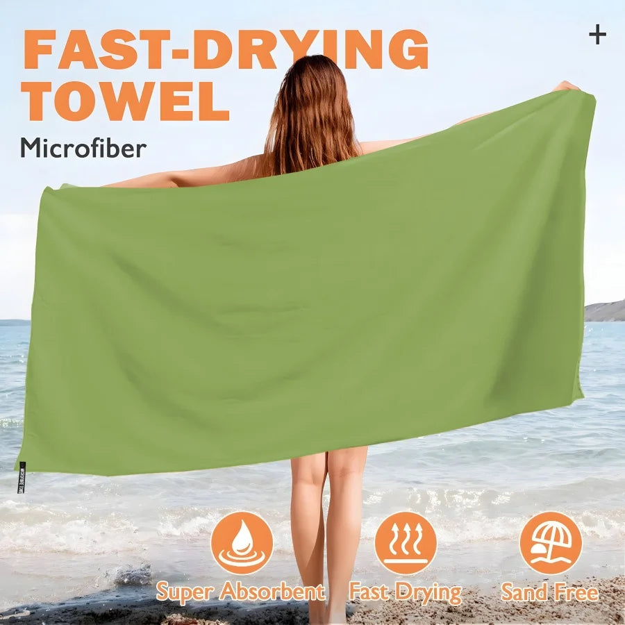 A woman holds up a large olive green microfiber quick-dry beach towel near the ocean water.