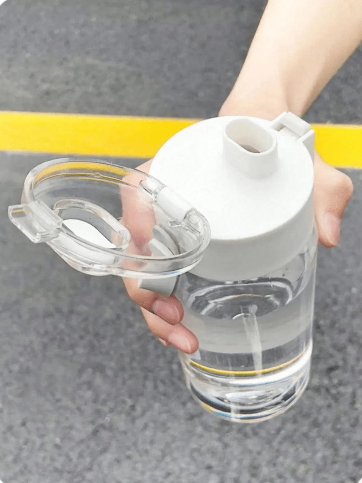 A hand holds a large capacity transparent sports water bottle with a white cap and straw, set against a gray background.