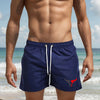A muscular man wearing navy blue quick-drying swim shorts with a red stylized bull logo on a beach.