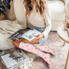 Eine Person trägt rosa Blumenmuster-Kompressionsstrümpfe und entspannt sich drinnen mit Büchern und einer weichen Decke.