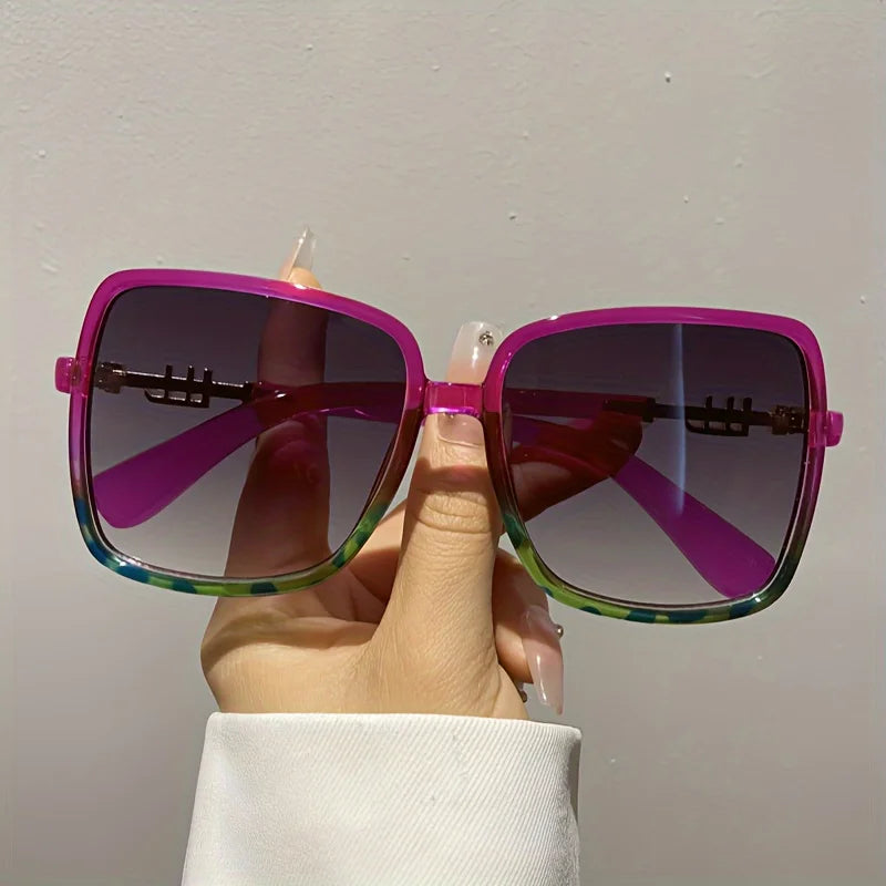 A hand holds a pair of retro two-tone gradient sunglasses with purple and green lenses against a light background.