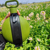 A hand holds a lime green collapsible silicone camping kettle with a black handle in a field of pink and white flowers.
