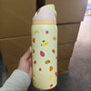 A light yellow insulated stainless steel sports water bottle with colorful fruit illustrations is held against a brown cardbo