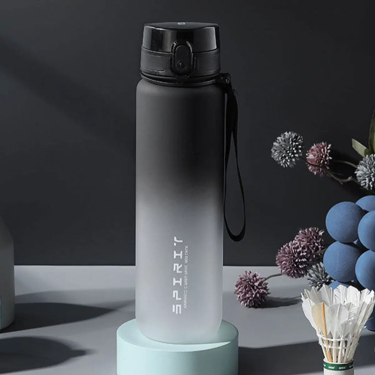 Eine hohe, matte graue isolierte Wasserflasche mit schwarzem Verschluss und Riemen wird auf einem hellblauen Ständer präsenti