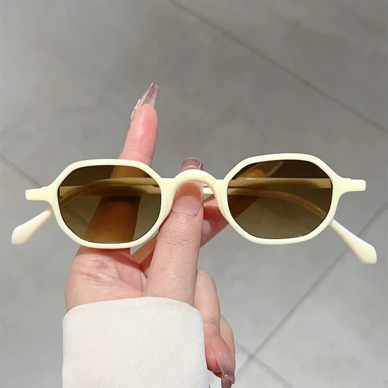 A hand holds a pair of vintage oval sunglasses with light beige frames and brown lenses.