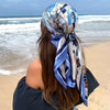 A woman with long brown hair wearing an elegant blue and silver satin square scarf tied around her hair by the ocean.