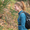A young woman with blonde hair wearing a blue jacket and a black backpack walks along a wooded path.