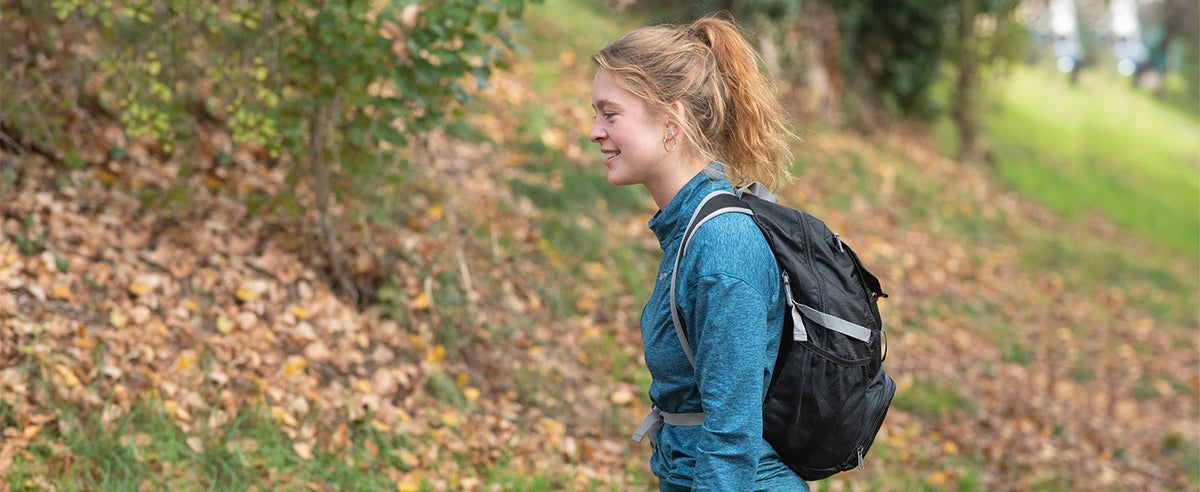 A young woman with blonde hair wearing a blue jacket and a black backpack walks along a wooded path.