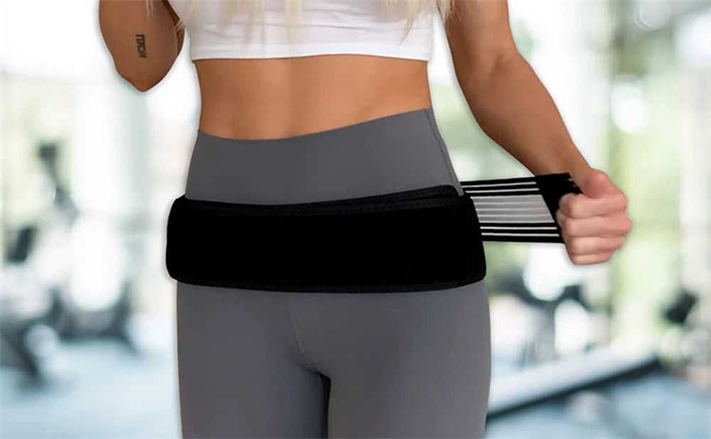 A woman wearing gray athletic wear is using a black adjustable pelvic support hip belt for lower back and SI joint relief.