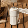 A white thermo mug with a digital temperature display sits on a wooden surface next to dried grasses.