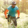 Ein Mann trägt ein türkises Sportshirt und orangefarbene Ärmel und fährt Fahrrad in einer sonnigen, grünen Umgebung.