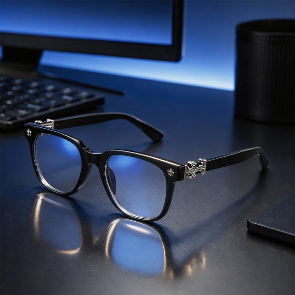 A pair of black square anti-blue light sunglasses resting on a dark surface next to a computer monitor.