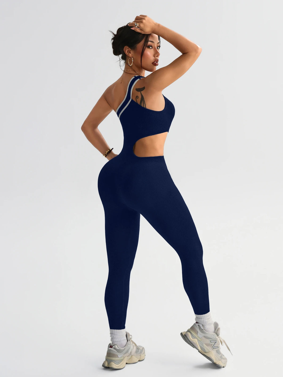 A woman wearing a navy blue seamless yoga jumpsuit with a cutout, posing against a light gray background.