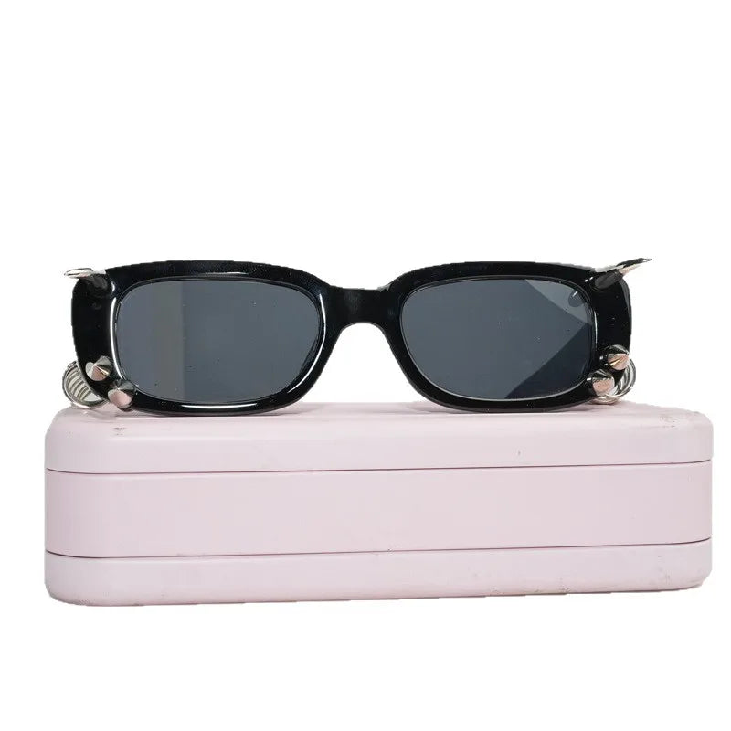 Black spiked rivet sunglasses resting on a pink rectangular case.