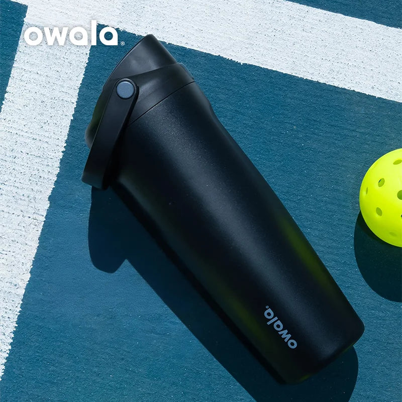 A black Owala insulated stainless steel travel mug resting on a blue surface next to a yellow tennis ball.
