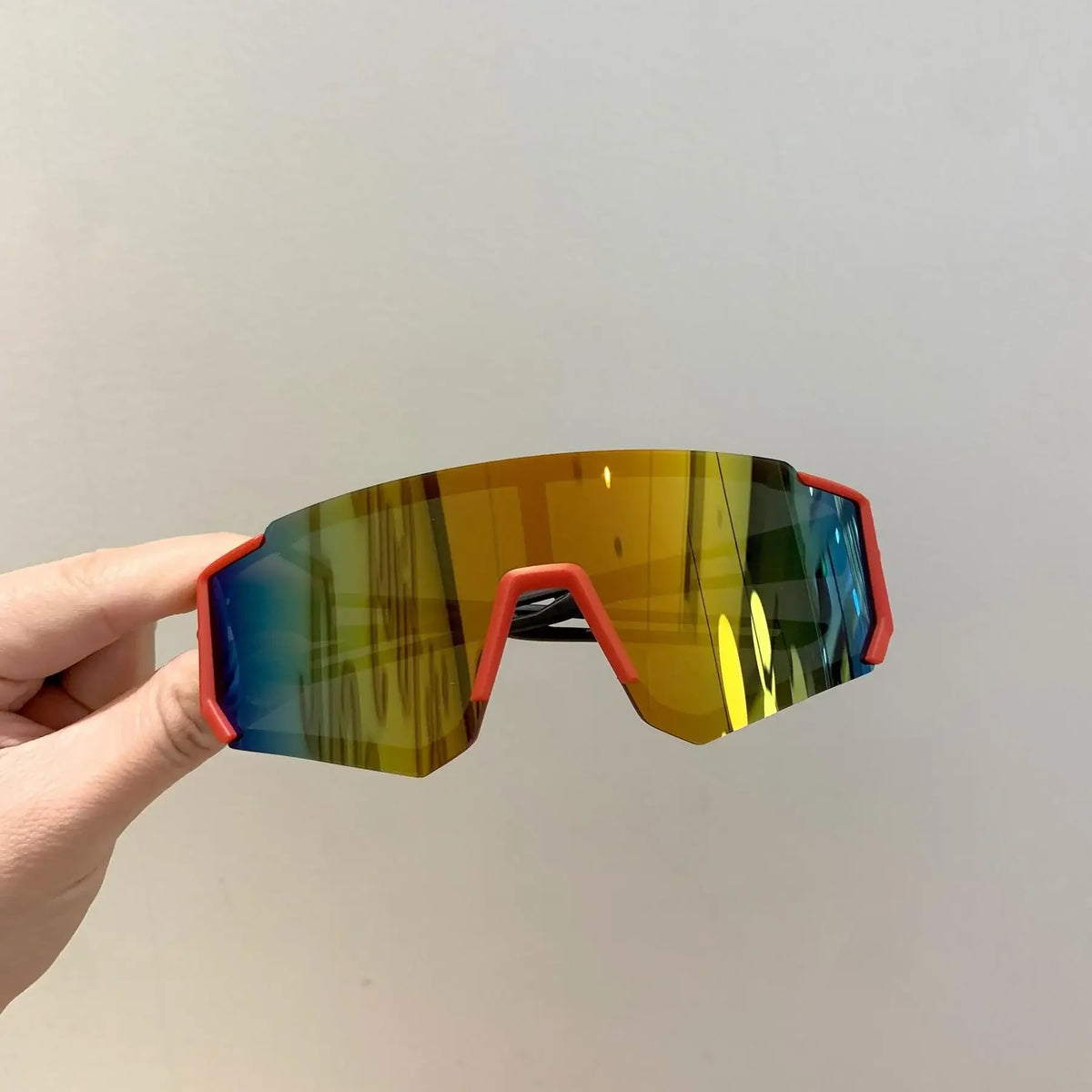 A hand holds a pair of orange-framed sports sunglasses with mirrored green and blue lenses.