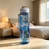 A clear blue water bottle with a strawberry bow design sits on a wooden table next to a bed.