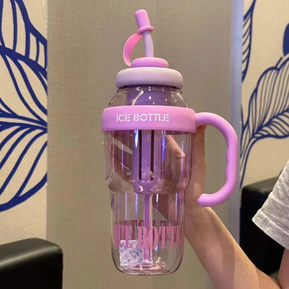 A hand holds a purple glass bottle with a pink lid and handle, demonstrating the large capacity portable plastic drinking cup