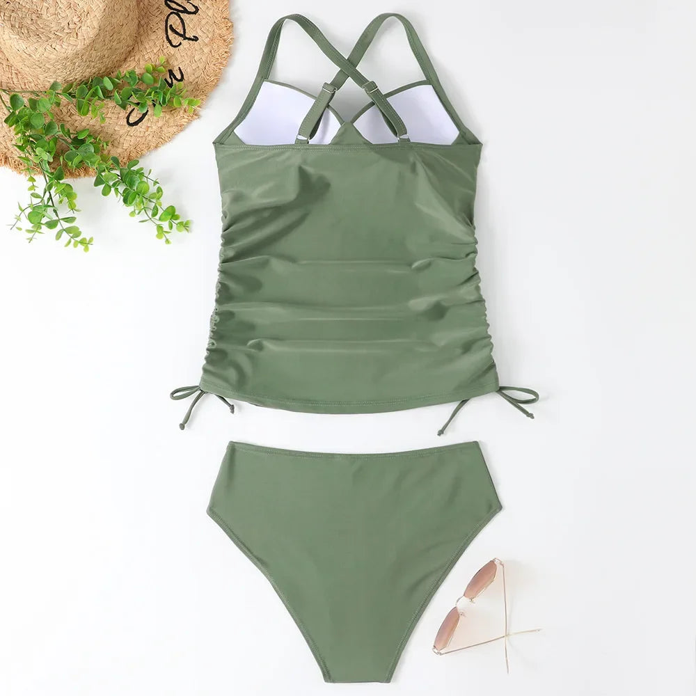 A sage green high-waisted two-piece swimsuit set laid out on a white surface with a straw hat nearby.