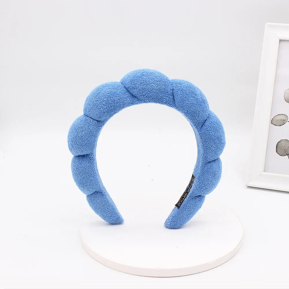 A light blue sponge terry cloth headband for hair styling displayed on a white circular stand.