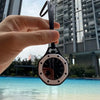 A hand holds an octagonal quartz pocket watch with a black strap, positioned above clear blue water.
