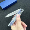 A hand holds a silver precision folding pocket knife with a blue and white patterned handle against a dark background.