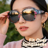 A woman models wrap-around sunglasses with floral patterns outdoors during sunset.