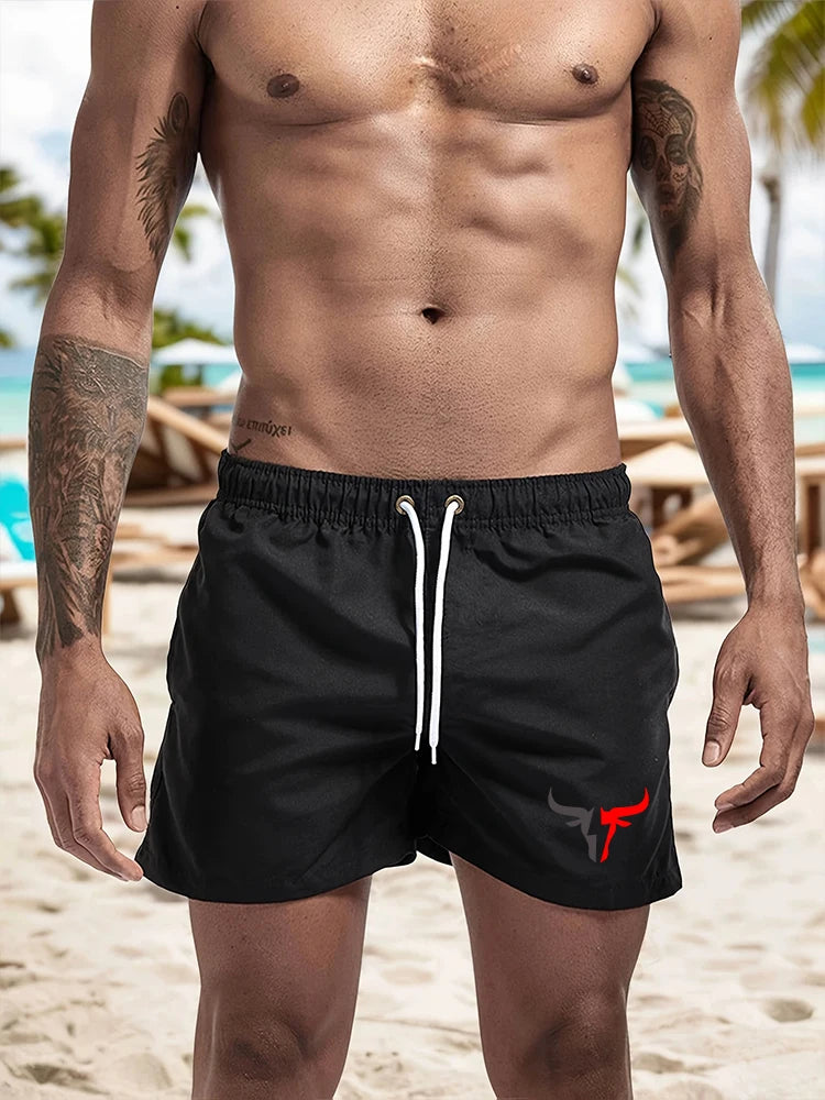A muscular man wearing black quick-drying swim shorts with a red bull emblem.