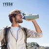 A man holds a light green insulated stainless steel water bottle outdoors against a blue sky.