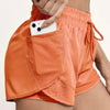 A hand is adjusting the mesh pocket on an orange athletic short featuring a cell phone.