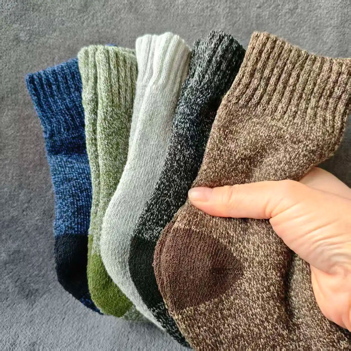 A hand holds a stack of three merino wool hiking socks in various shades of blue, green, and brown.
