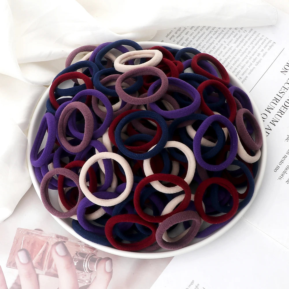 A pile of 50 pieces of mixed color elastic hair bands in various shades of purple, blue, and maroon on a white plate.