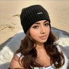 A woman wearing a black knitted winter headwrap and a white top, posing outdoors on sand.