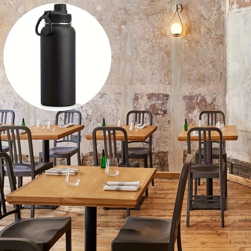 A black insulated stainless steel vacuum water bottle standing on a wooden table in a rustic indoor setting.