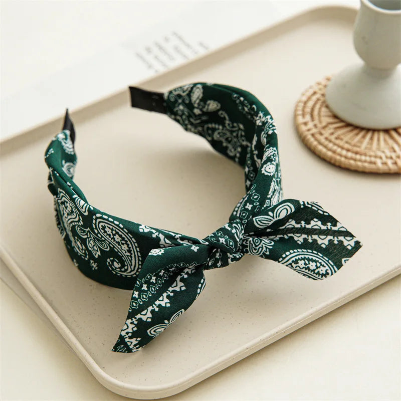 A dark green paisley bandana headband tied in a bow, displayed on a light tray.
