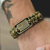 A close-up of a tactical paracord survival bracelet with a bronze American flag charm worn on a wrist.
