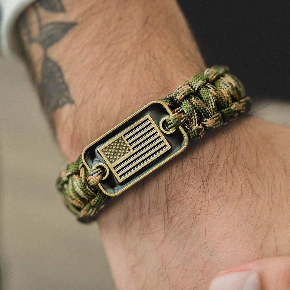 A close-up of a tactical paracord survival bracelet with a bronze American flag charm worn on a wrist.