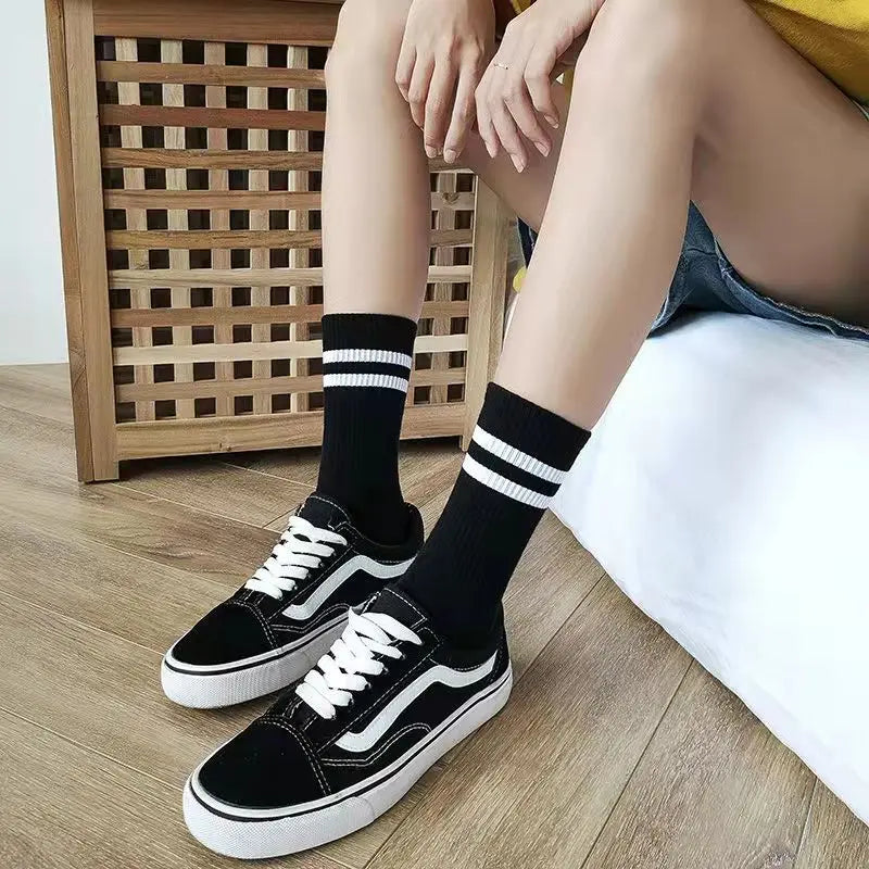 A pair of black and white athletic shoes with black socks are shown resting on a wooden floor next to a white pillow.