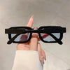 A hand holds a pair of black rectangular sunglasses with thin arms against a light background.