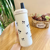Un tumbler isotherme blanc avec un couvercle à paille noire et de petits nœuds noirs est tenu contre une surface en bois.
