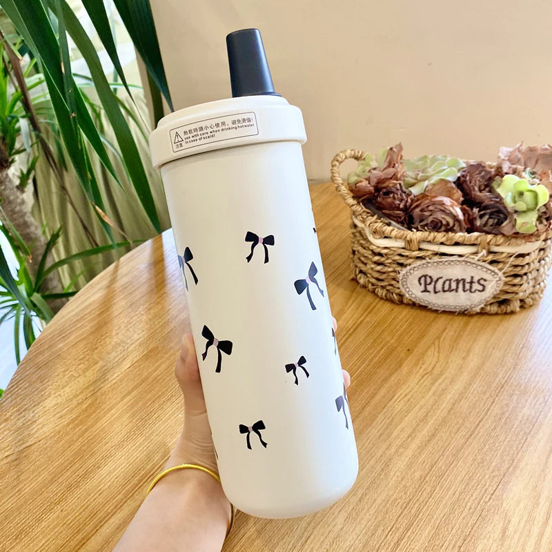 Un tumbler isotherme blanc avec un couvercle à paille noire et de petits nœuds noirs est tenu contre une surface en bois.