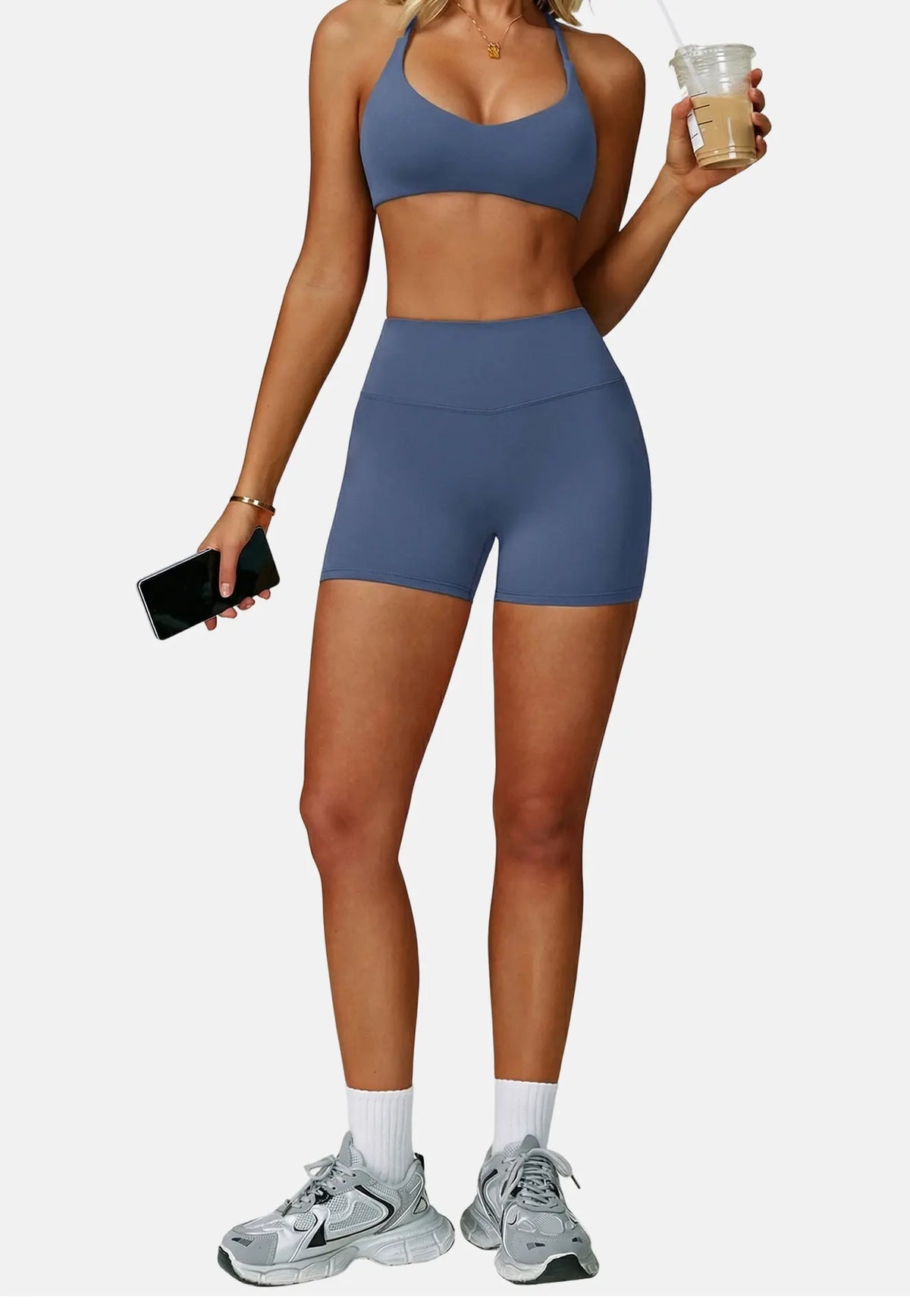 A woman wearing a navy blue high-support sports bra and matching shorts, holding a drink and a phone.