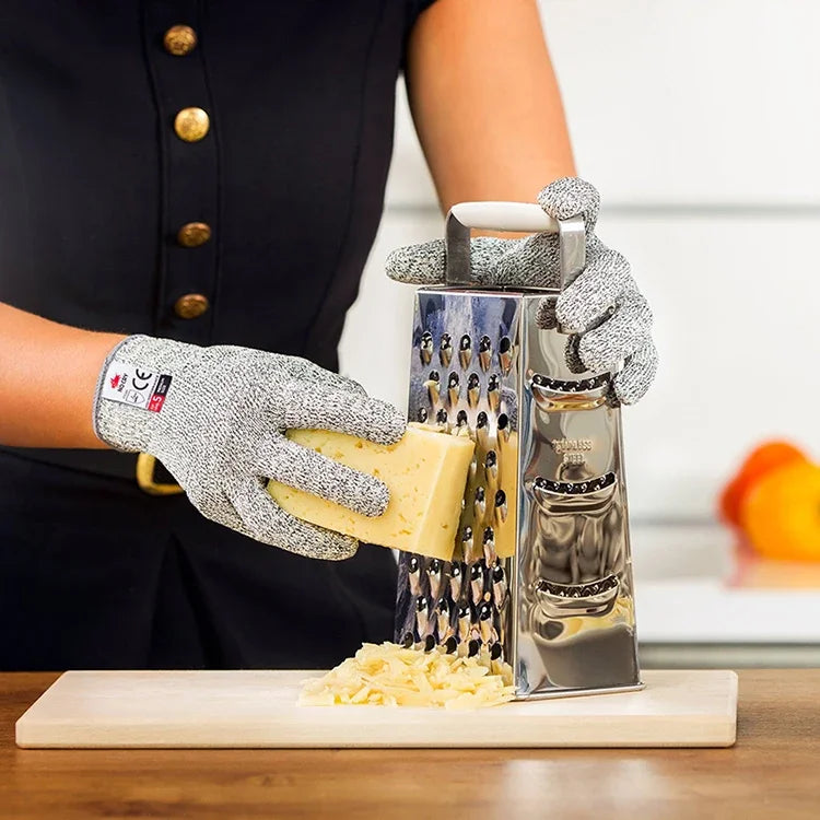 A hand wearing knitted protection gloves is holding a block of cheese over a wooden board.