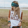 A blonde woman wearing a teal patterned headband and a white t-shirt with brown text stands in an open field.