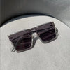 A pair of dark purple tinted square sunglasses with a thin silver frame resting on a light gray surface.