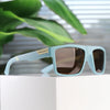 Light blue polarized square sunglasses resting on a white surface with green foliage in the background.