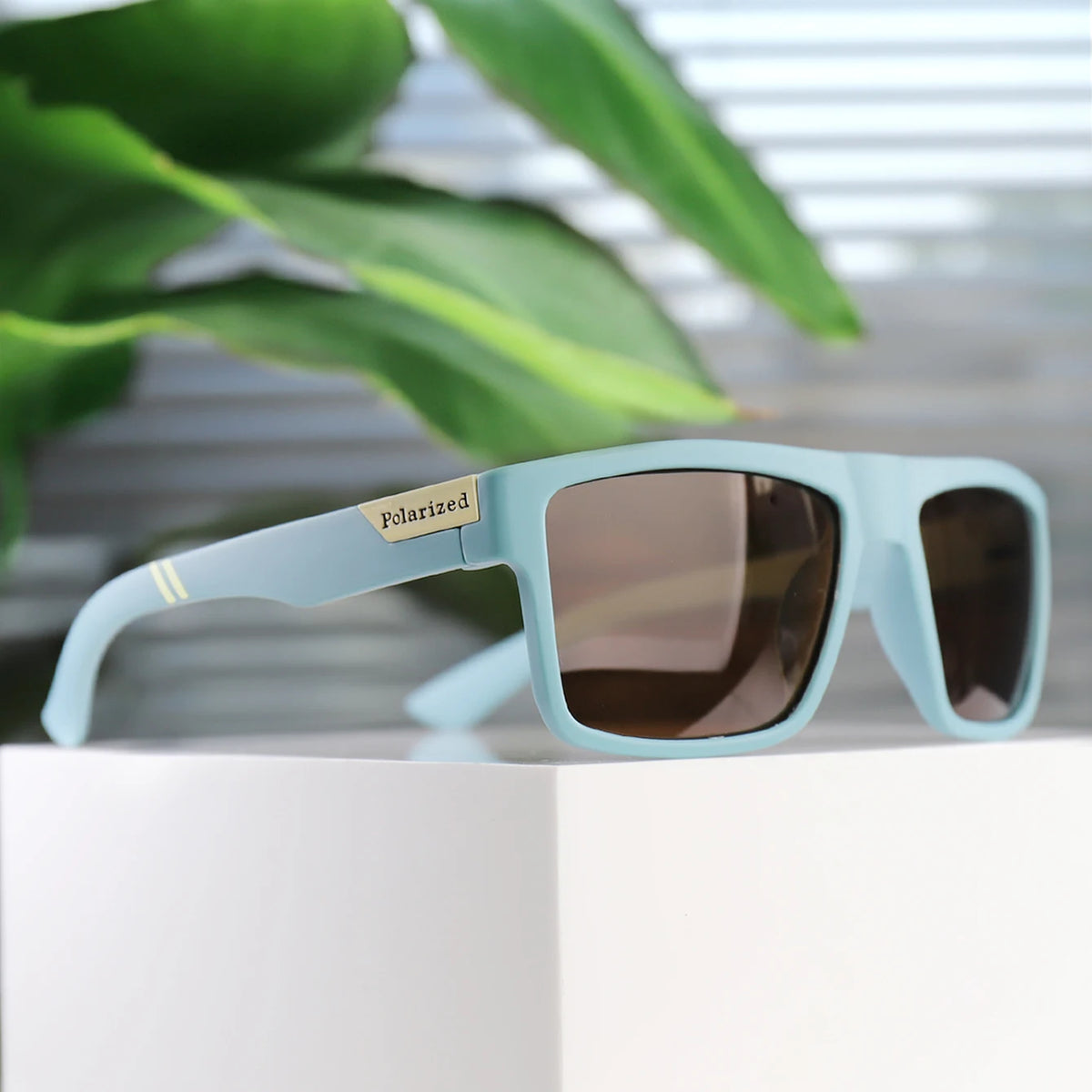Light blue polarized square sunglasses resting on a white surface with green foliage in the background.